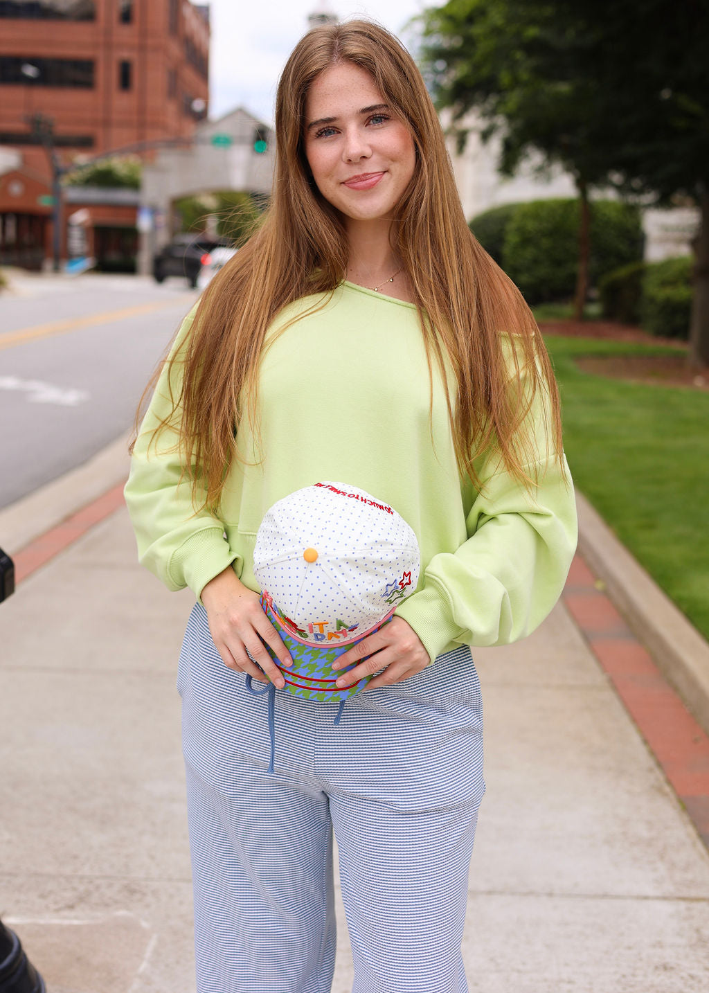 A woman standing on a city sidewalk wearing a light lime green long-sleeve top and light blue pinstriped pants, holding a white polka-dot bucket hat.