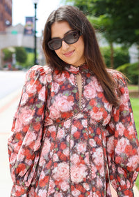 A woman wearing a pink and red floral long-sleeve mini dress with puff sleeves, styled with dark sunglasses and a brown handbag, standing on an outdoor sidewalk.