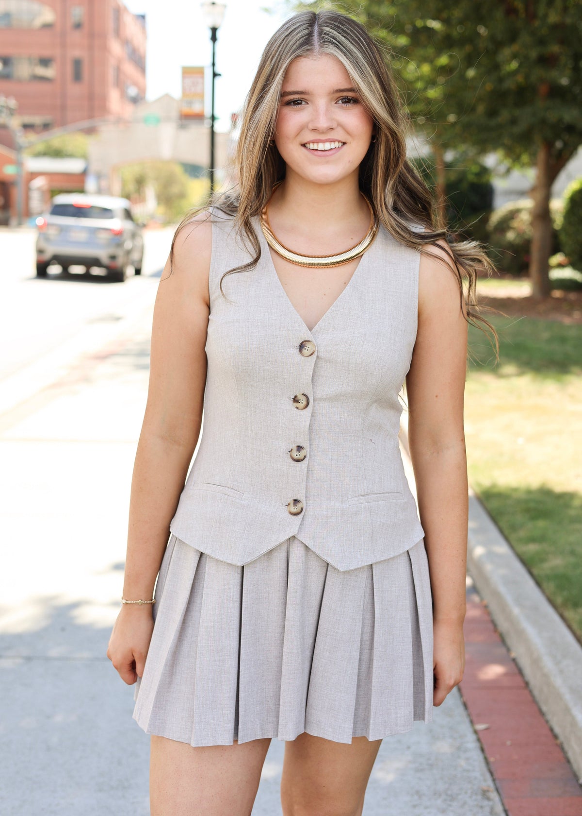 A young woman smiling on a city sidewalk, wearing a light beige sleeveless vest and matching pleated mini skirt set with gold buttons and a gold necklace.