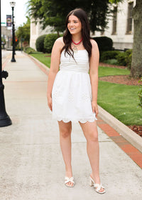 Woman in a white dress standing on a sidewalk with greenery and a building in the background