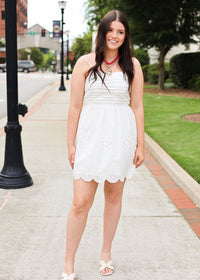 Woman in a white dress standing on a sidewalk with greenery and buildings in the background