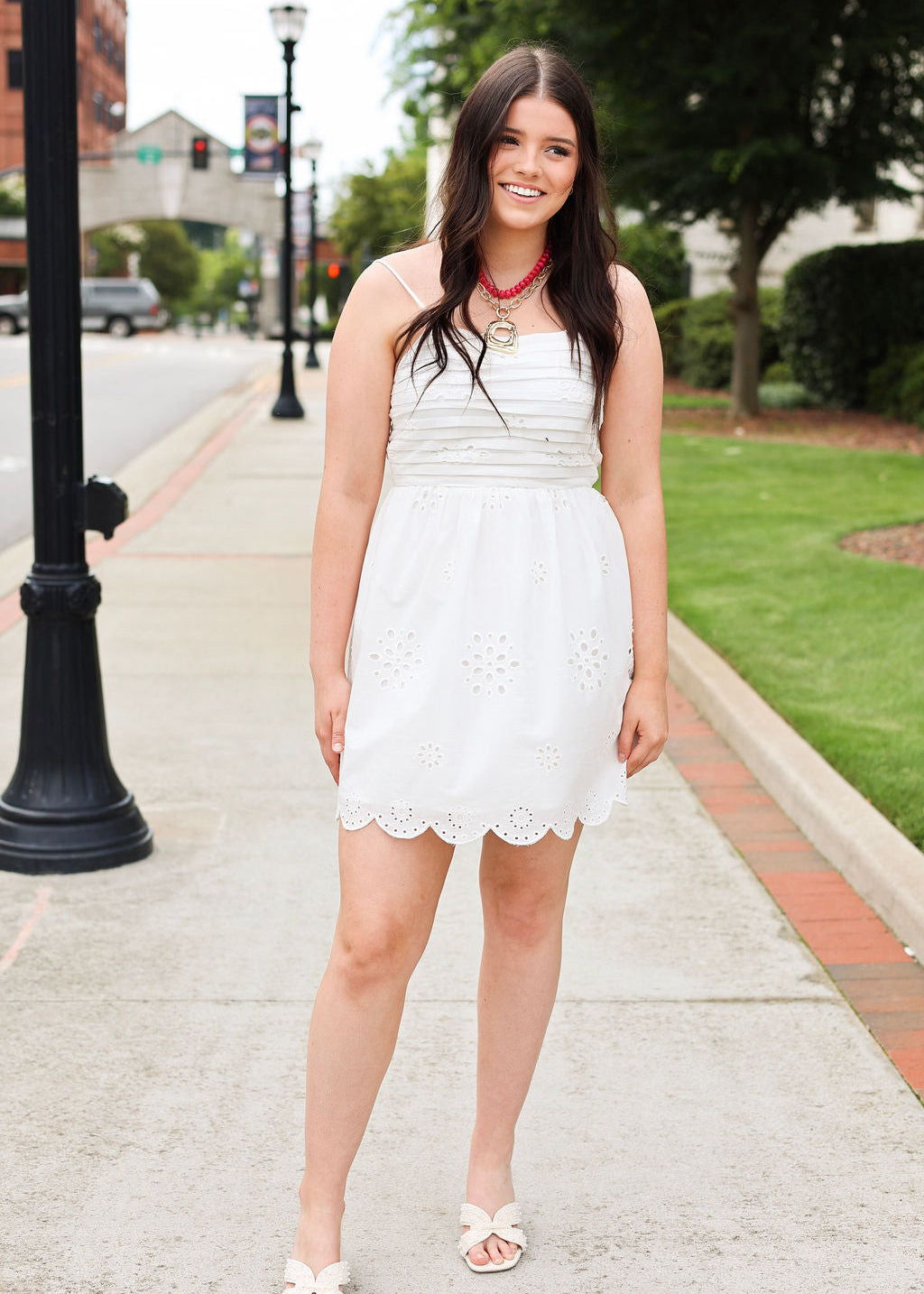 Woman in a white dress standing on a sidewalk with greenery and buildings in the background