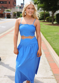 Woman in a blue dress standing on a sidewalk with greenery and buildings in the background