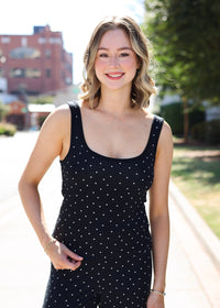 A woman wearing a black sleeveless jumpsuit with small white polka dots, standing outdoors on a sunny day with trees and a brick building blurred in the background.