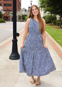 Woman in a blue floral dress standing on a sidewalk with buildings and greenery in the background