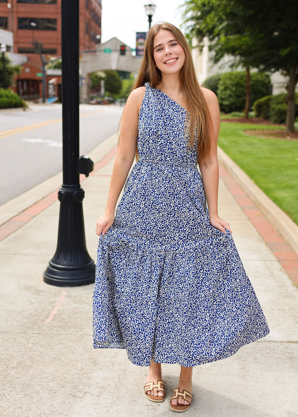 Woman in a blue floral dress standing on a sidewalk with buildings and greenery in the background