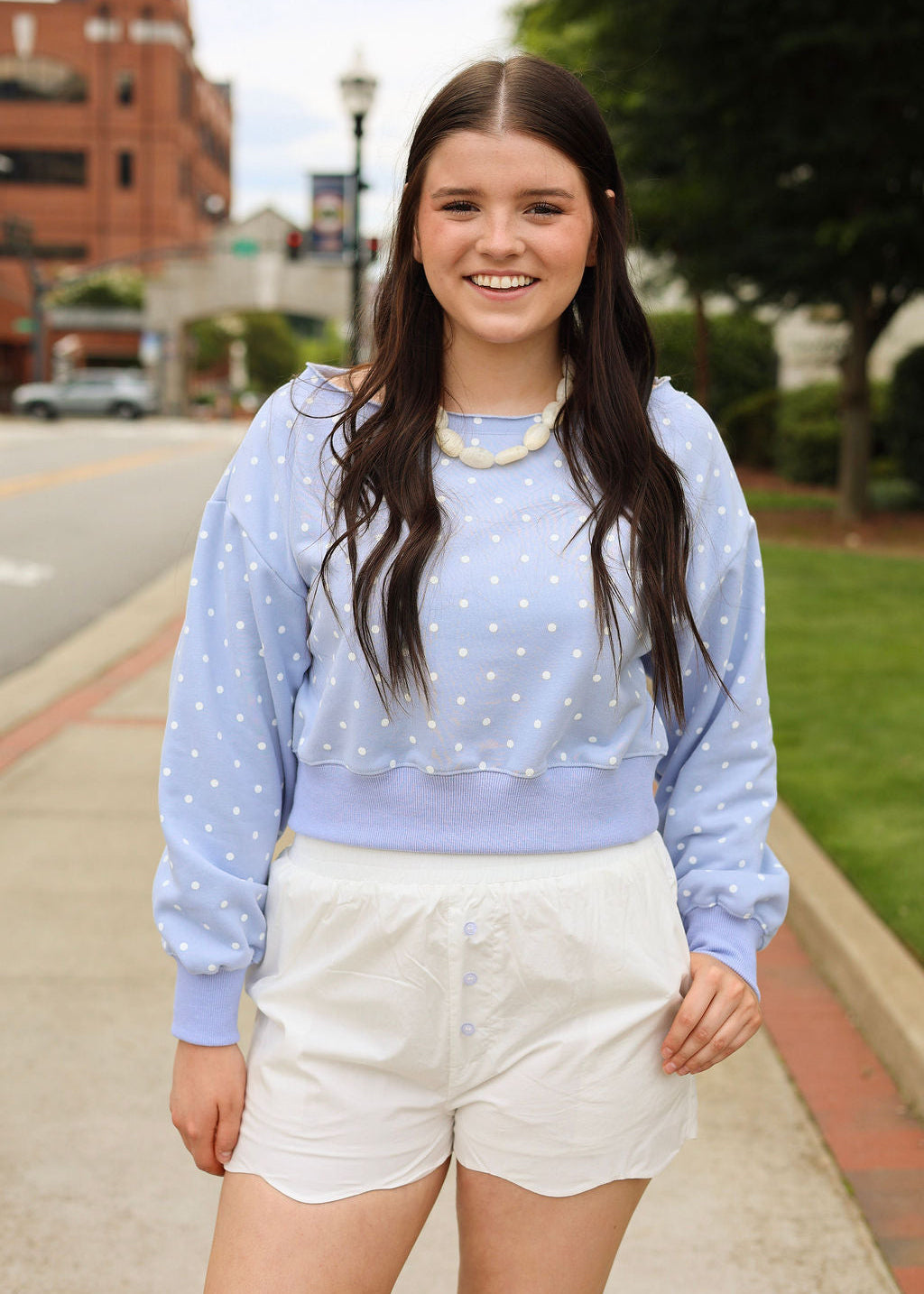A young woman smiling outdoors on a sidewalk, wearing a light blue long-sleeve cropped sweatshirt with white polka dots and white shorts.