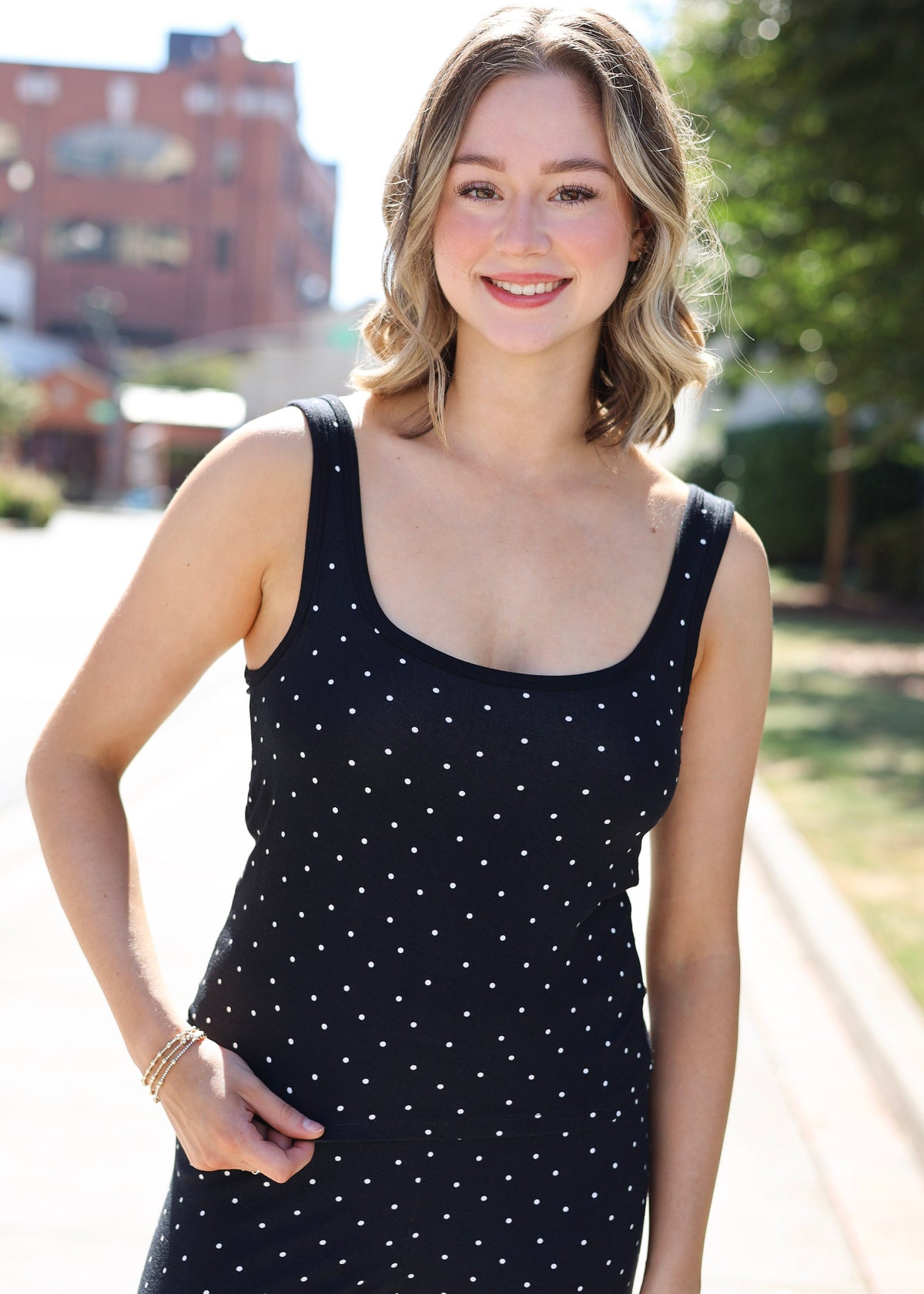 A woman wearing a black sleeveless jumpsuit with small white polka dots, standing outdoors on a sunny day with a blurred background of trees and buildings.