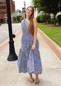 Woman in a blue floral dress standing on a sidewalk with a street and greenery in the background