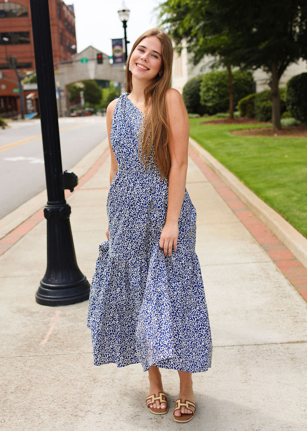 Woman in a blue floral dress standing on a sidewalk with a street and greenery in the background