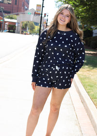 A woman modeling a navy blue long-sleeve sweatshirt and matching shorts set with a white floral pattern, standing on a sunny outdoor sidewalk.
