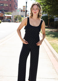 A woman modeling a black sleeveless jumpsuit with white polka dots on a sunny city sidewalk.