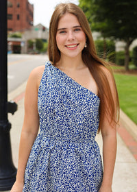 Woman wearing a blue floral dress standing outdoors on a sidewalk.