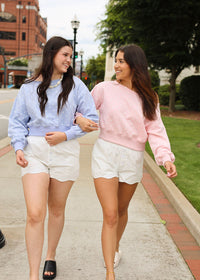 Two women walking arm-in-arm on an outdoor sidewalk. The woman on the left wears a light blue long-sleeve top with white polka dots and white shorts, while the woman on the right wears a light pink long-sleeve top and white shorts.
