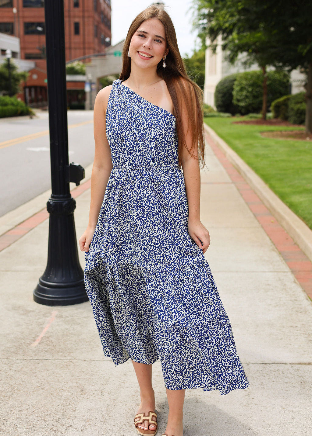 Woman in a blue floral dress standing on a sidewalk with a building and trees in the background.