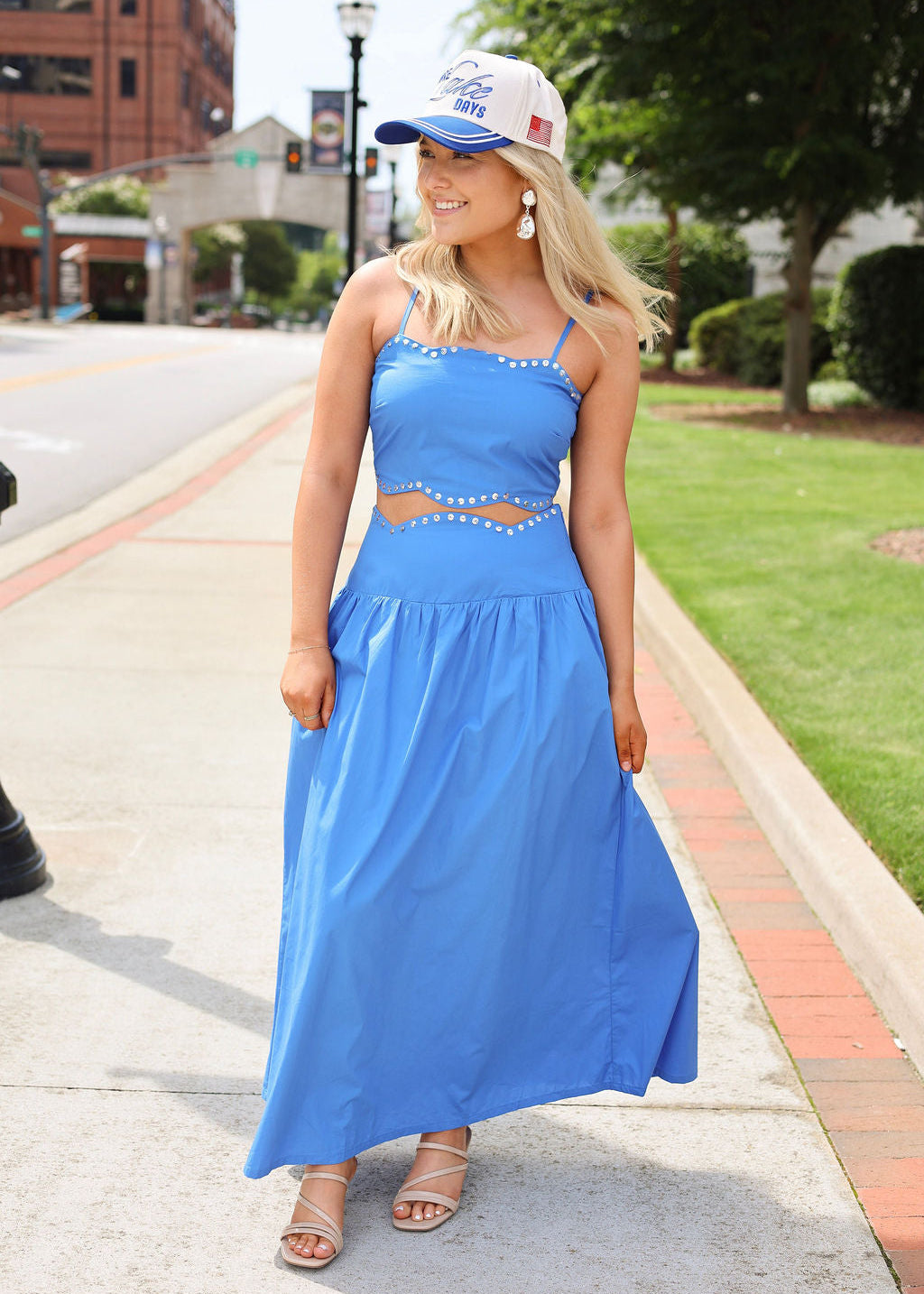 Woman in a blue dress and cap standing on a sidewalk with greenery and buildings in the background