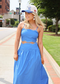 Woman in a blue dress and cap standing on a sidewalk with greenery and buildings in the background