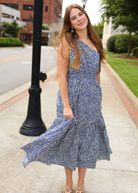 Woman in a blue floral dress standing on a sidewalk with buildings and greenery in the background