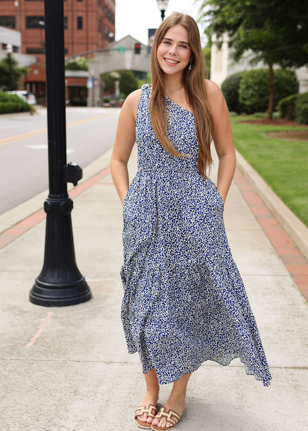 Woman in a blue floral dress standing on a sidewalk with buildings and greenery in the background
