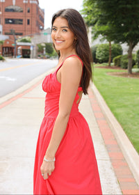 Woman in a red dress standing on a sidewalk with buildings and greenery in the background