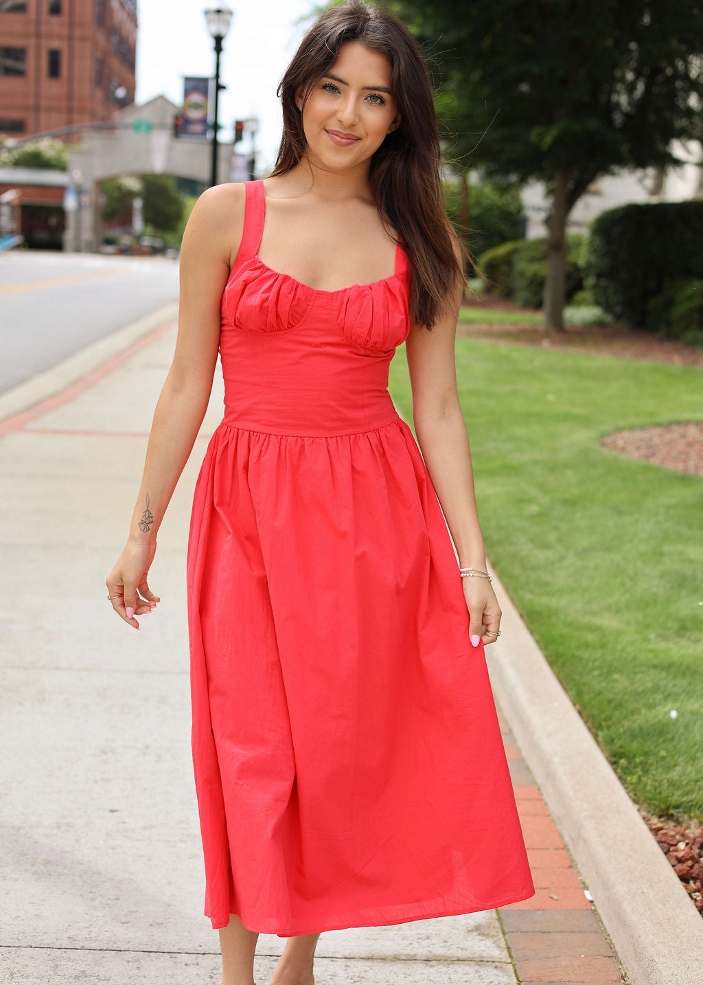 Woman in a red dress standing on a sidewalk with greenery in the background