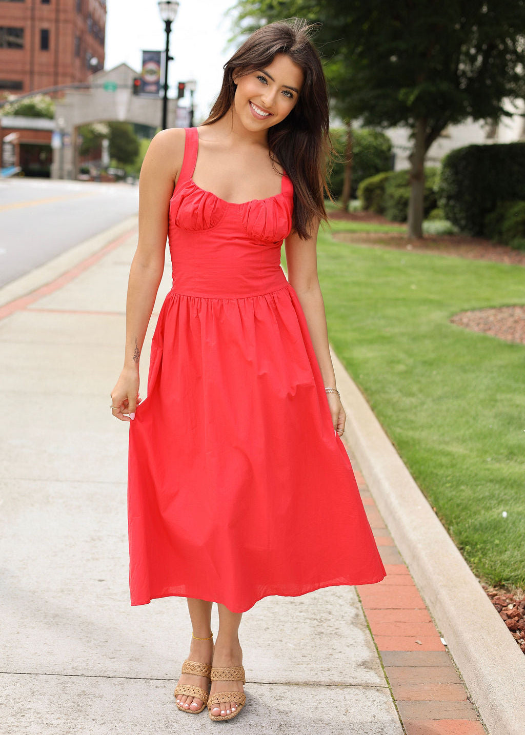 Woman in a red dress standing on a sidewalk with greenery and buildings in the background