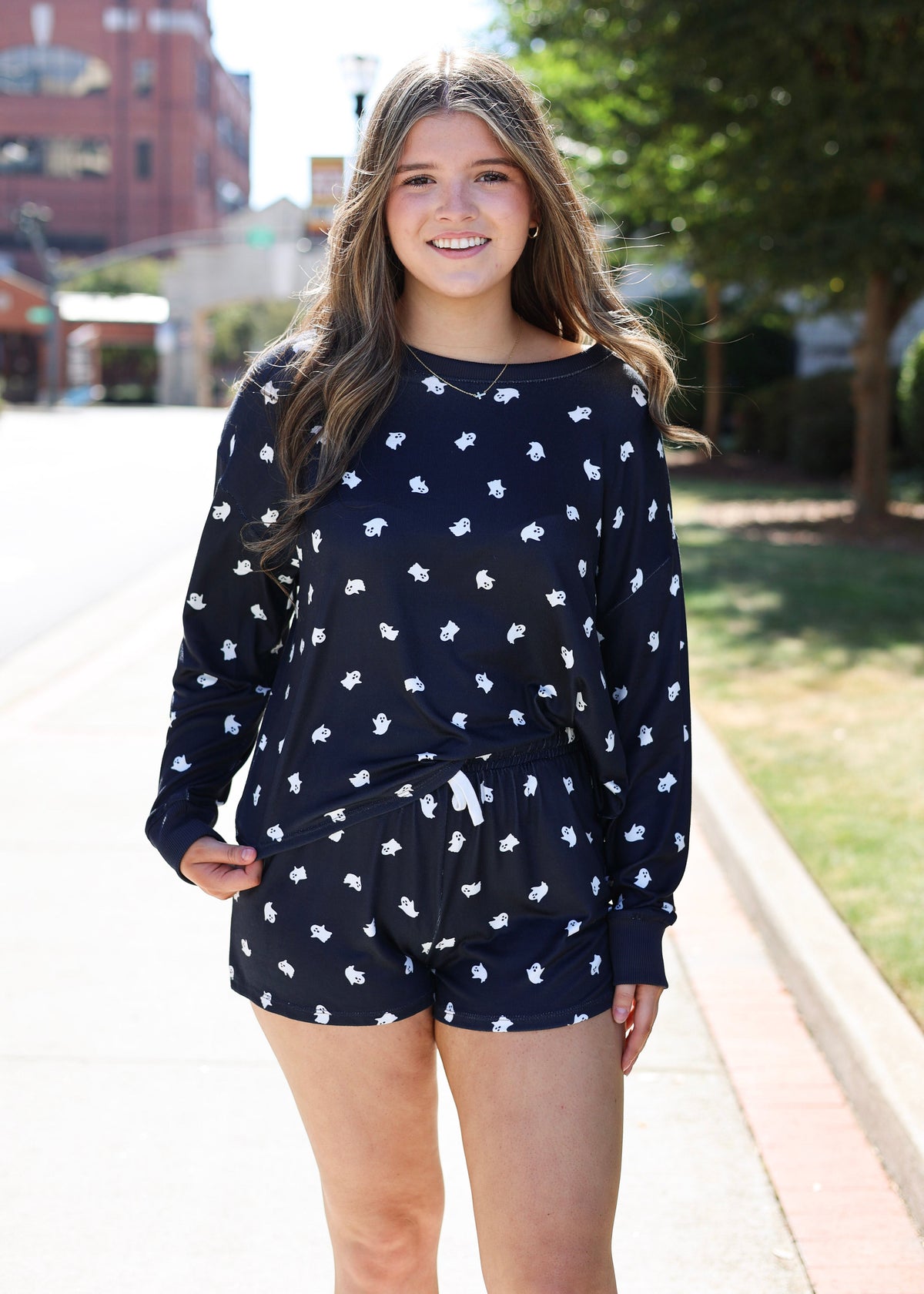 A young woman smiling outdoors on a sunny day, wearing a navy blue long-sleeve top and matching shorts set with a white floral pattern.