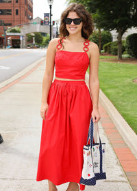 Woman in a red dress with decorative straps standing on a sidewalk.