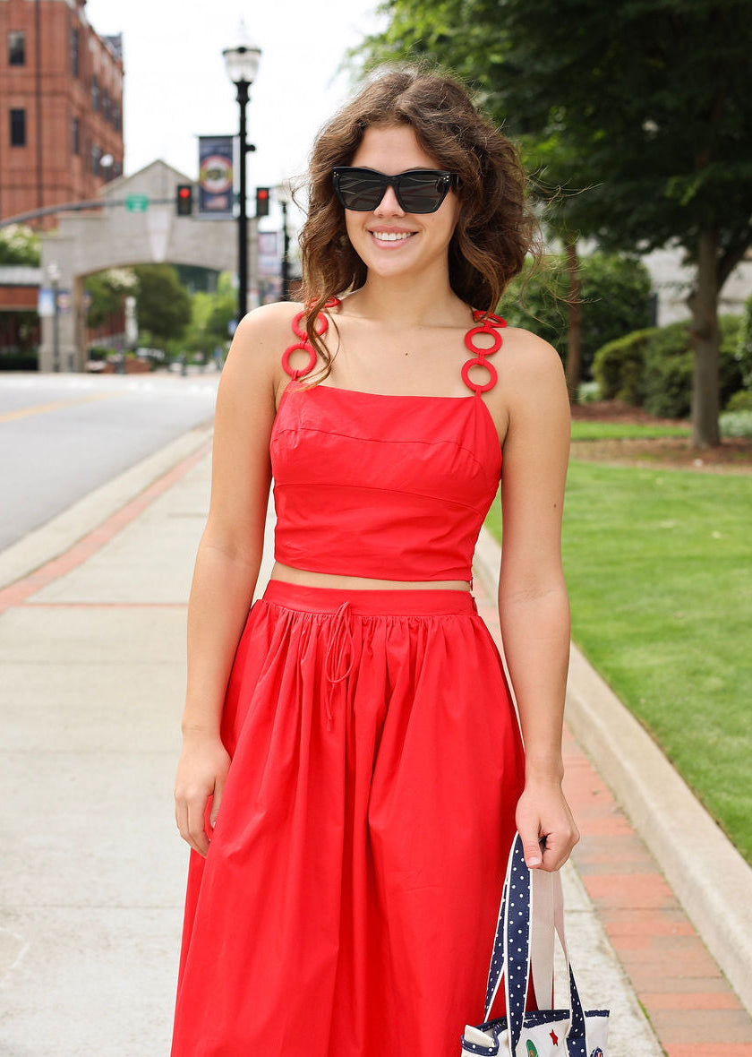 Woman in a red dress with decorative straps standing on a sidewalk.