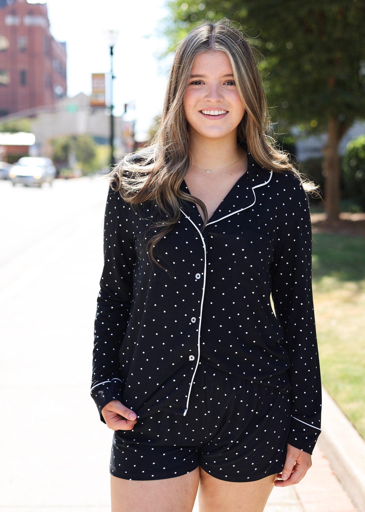 A smiling young woman stands outdoors on a sidewalk wearing a matching black pajama set with small white polka dots and white piping trim.