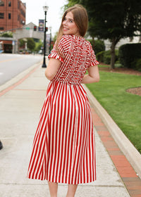 Woman in a red and white striped dress standing on a sidewalk with buildings and trees in the background.