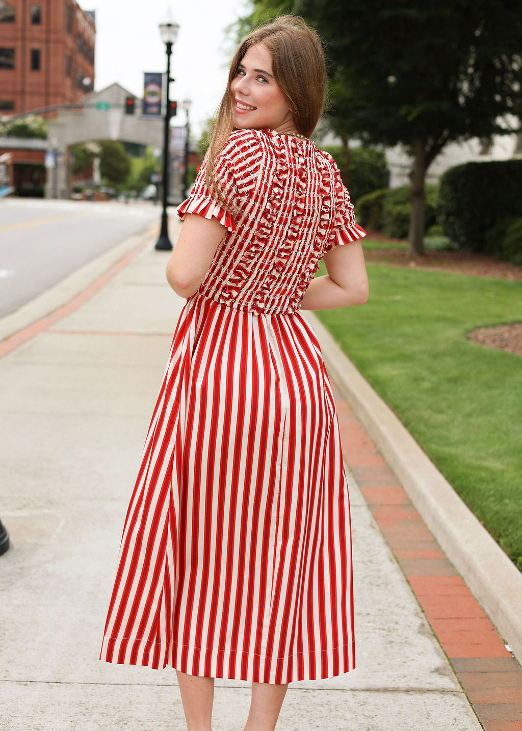 Woman in a red and white striped dress standing on a sidewalk with buildings and trees in the background.