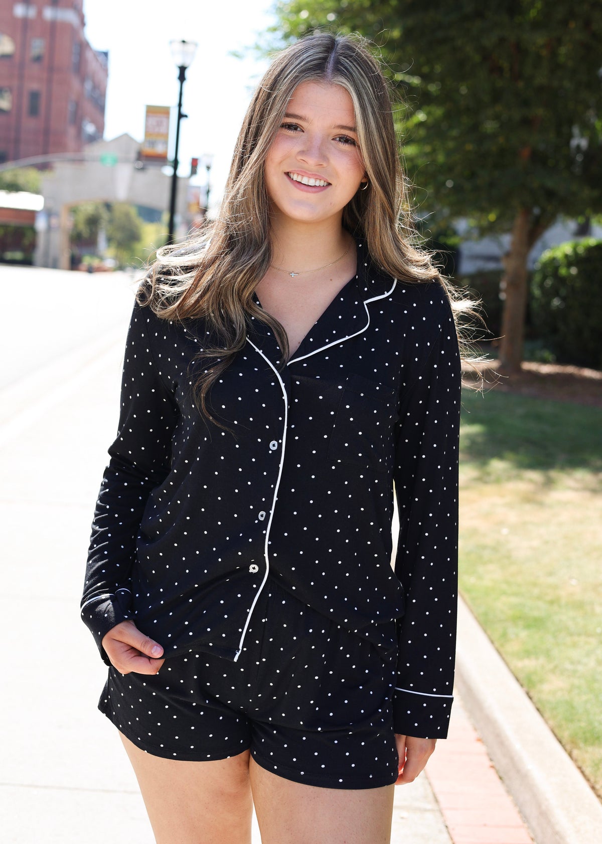 A woman wearing a black long-sleeve pajama shirt and matching shorts with small white polka dots and white piping, standing outdoors on a sunny day.