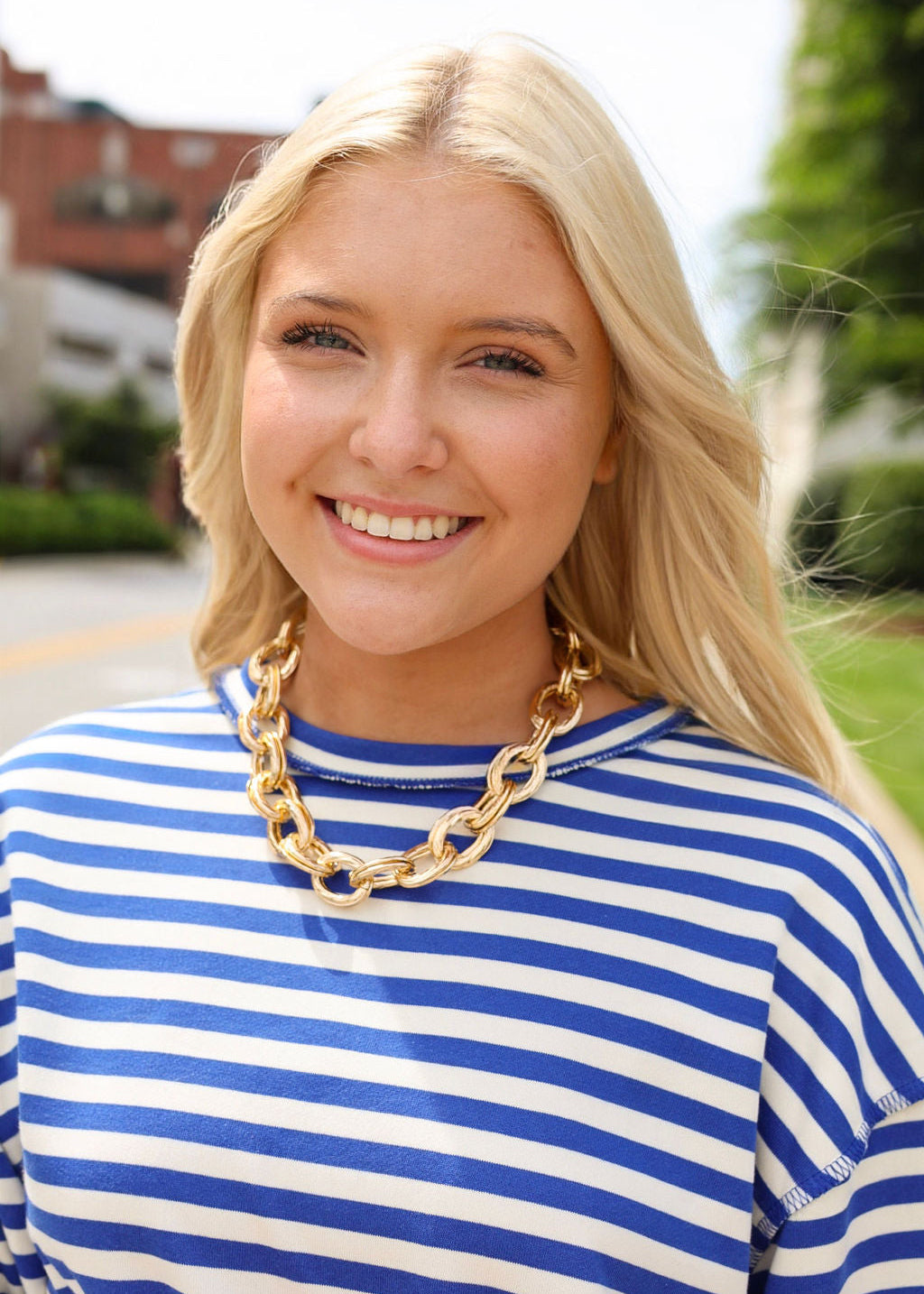 Woman wearing a blue and white striped shirt with a gold chain necklace outdoors.
