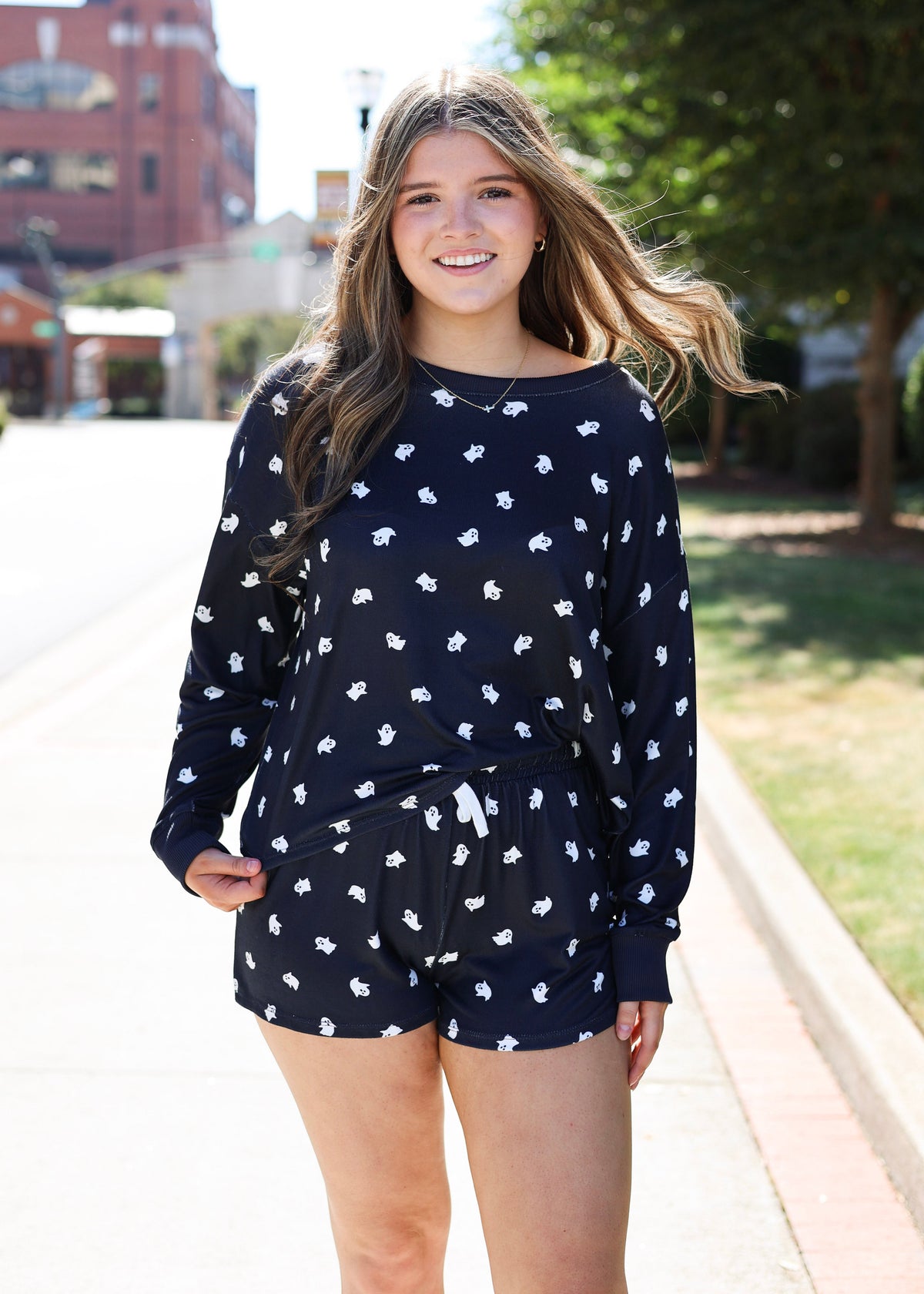 A young woman smiling on a sunny outdoor sidewalk, wearing a matching navy blue lounge set with a white dachshund print. The set includes a long-sleeve top and shorts with a white drawstring.