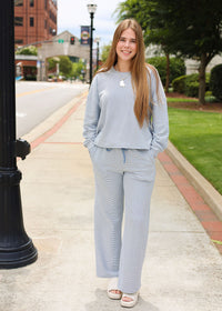 A young woman stands on a city sidewalk wearing a light blue-gray matching lounge set consisting of a cropped long-sleeve top and wide-leg pants, paired with white slide sandals.