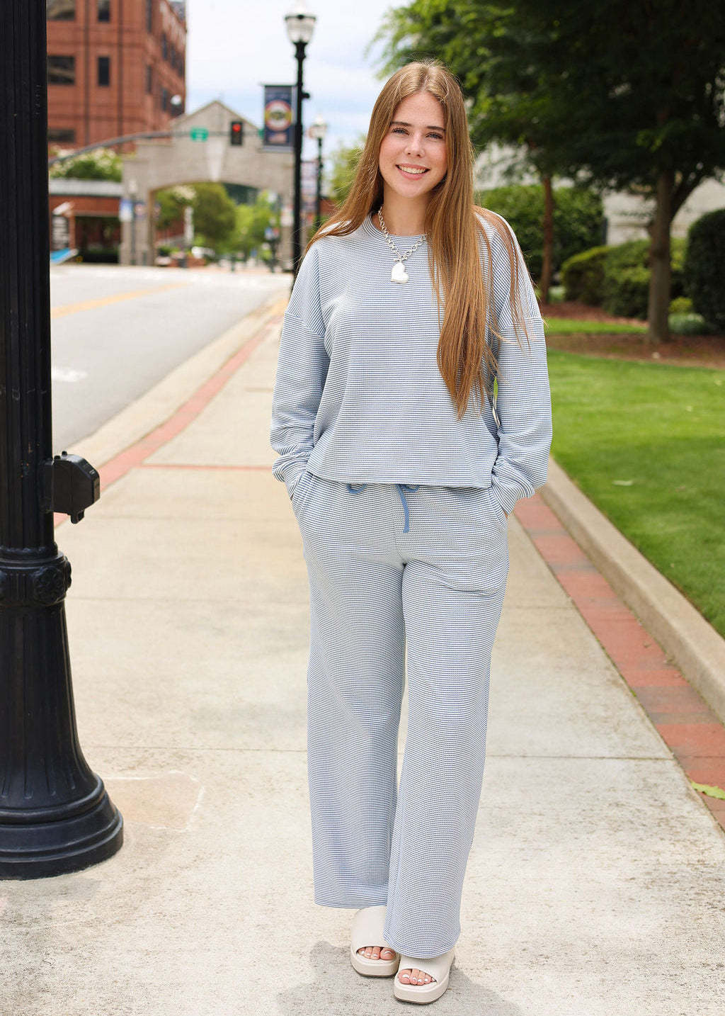 A young woman stands on a city sidewalk wearing a light blue-gray matching lounge set consisting of a cropped long-sleeve top and wide-leg pants, paired with white slide sandals.