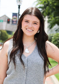 A young woman stands on a sunny sidewalk wearing a gray sleeveless knit vest and dark charcoal gray ruffled shorts, with trees and a brick building in the background.