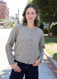 A woman wearing a light gray textured knit cardigan with white buttons and dark navy pants, standing outdoors on a sidewalk with trees and a street in the background.