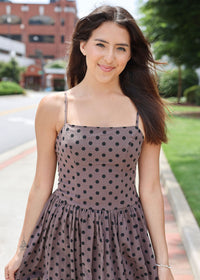 A woman wearing a brownish-gray spaghetti strap dress with black polka dots, standing outdoors on a sidewalk with trees and a brick building in the background.