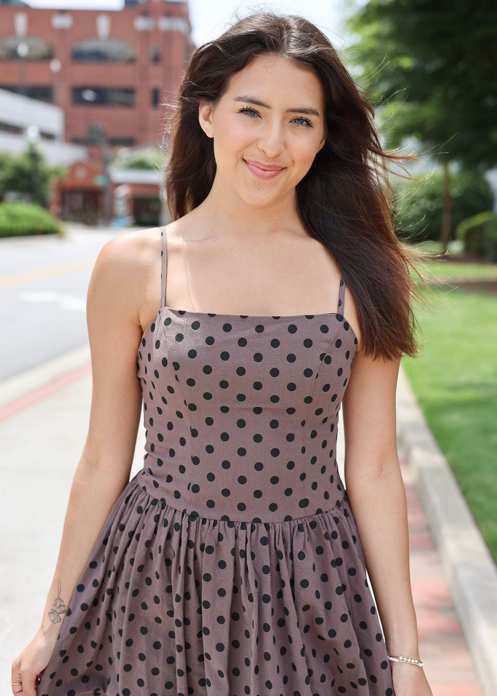 A woman wearing a brownish-gray spaghetti strap dress with black polka dots, standing outdoors on a sidewalk with trees and a brick building in the background.