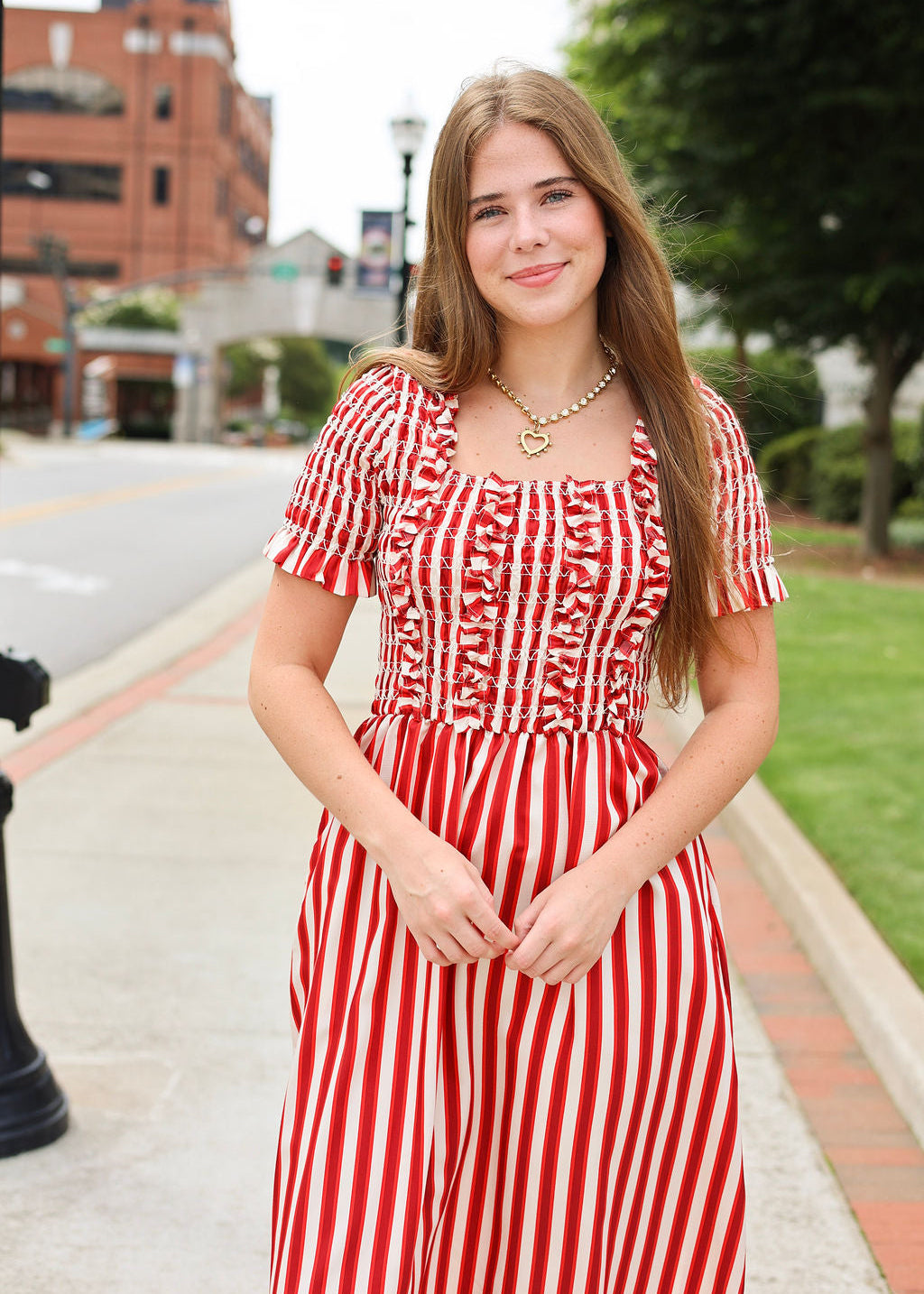 Woman in a red and white striped dress standing on a sidewalk with buildings and trees in the background.