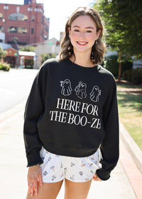 A woman standing outdoors on a sidewalk wearing a black crewneck sweatshirt with white ghost graphics and the text 'HERE FOR THE BOO-ZE', paired with white shorts featuring a small ghost pattern.