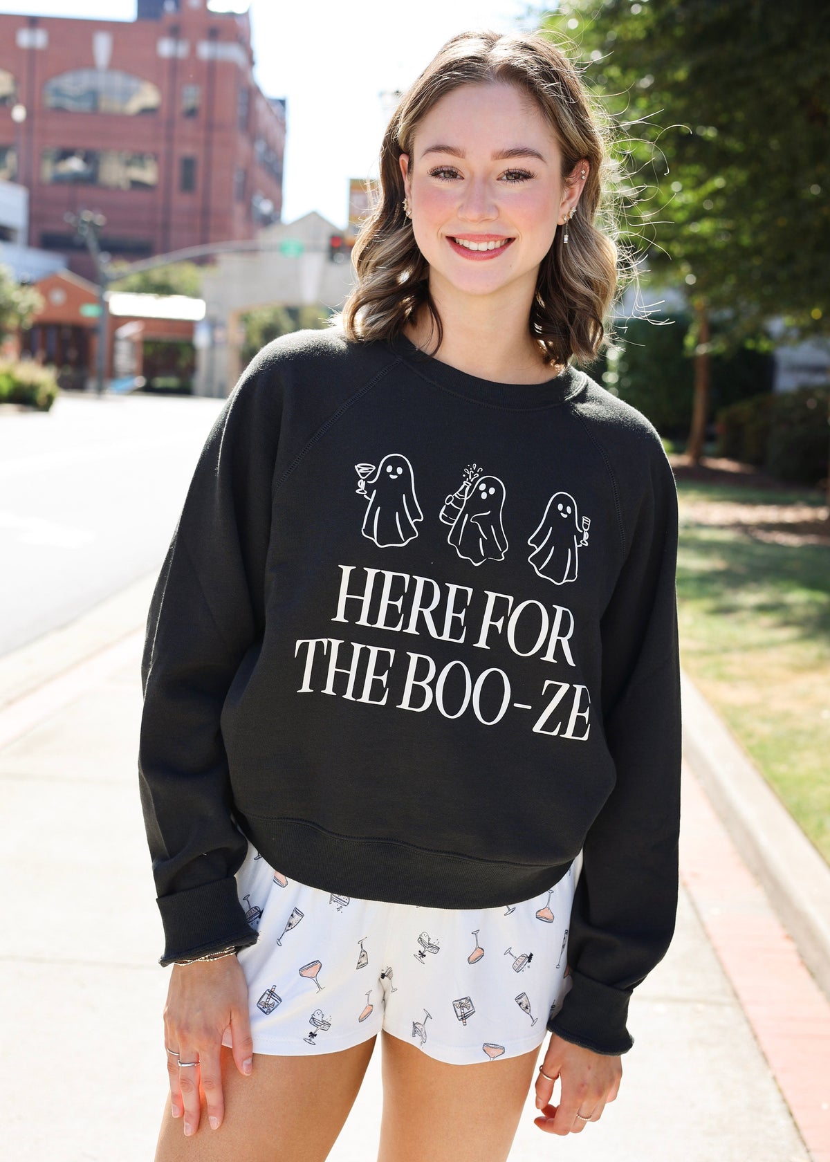 A woman standing outdoors on a sidewalk wearing a black crewneck sweatshirt with white ghost illustrations and the text 'HERE FOR THE BOO-ZE', paired with white shorts featuring small spooky prints.