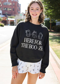 A woman standing outdoors on a sidewalk wearing a black crewneck sweatshirt with white ghost illustrations and the text 'HERE FOR THE BOO-ZE', paired with white shorts featuring small spooky prints.