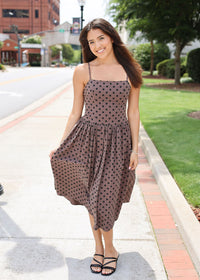 A woman wearing a brown spaghetti-strap midi dress with a black polka dot pattern, walking on a paved sidewalk outdoors.
