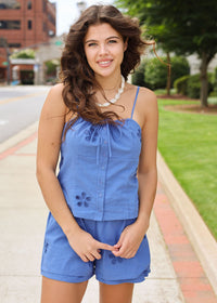 Woman wearing a blue romper standing on a sidewalk with greenery and buildings in the background
