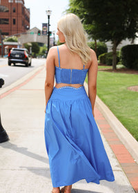 Woman in a blue dress walking on a sidewalk with buildings and greenery in the background