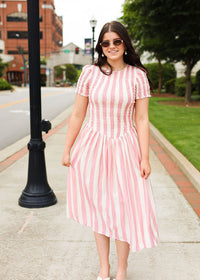 Woman in a pink and white striped dress standing on a sidewalk with buildings and trees in the background.