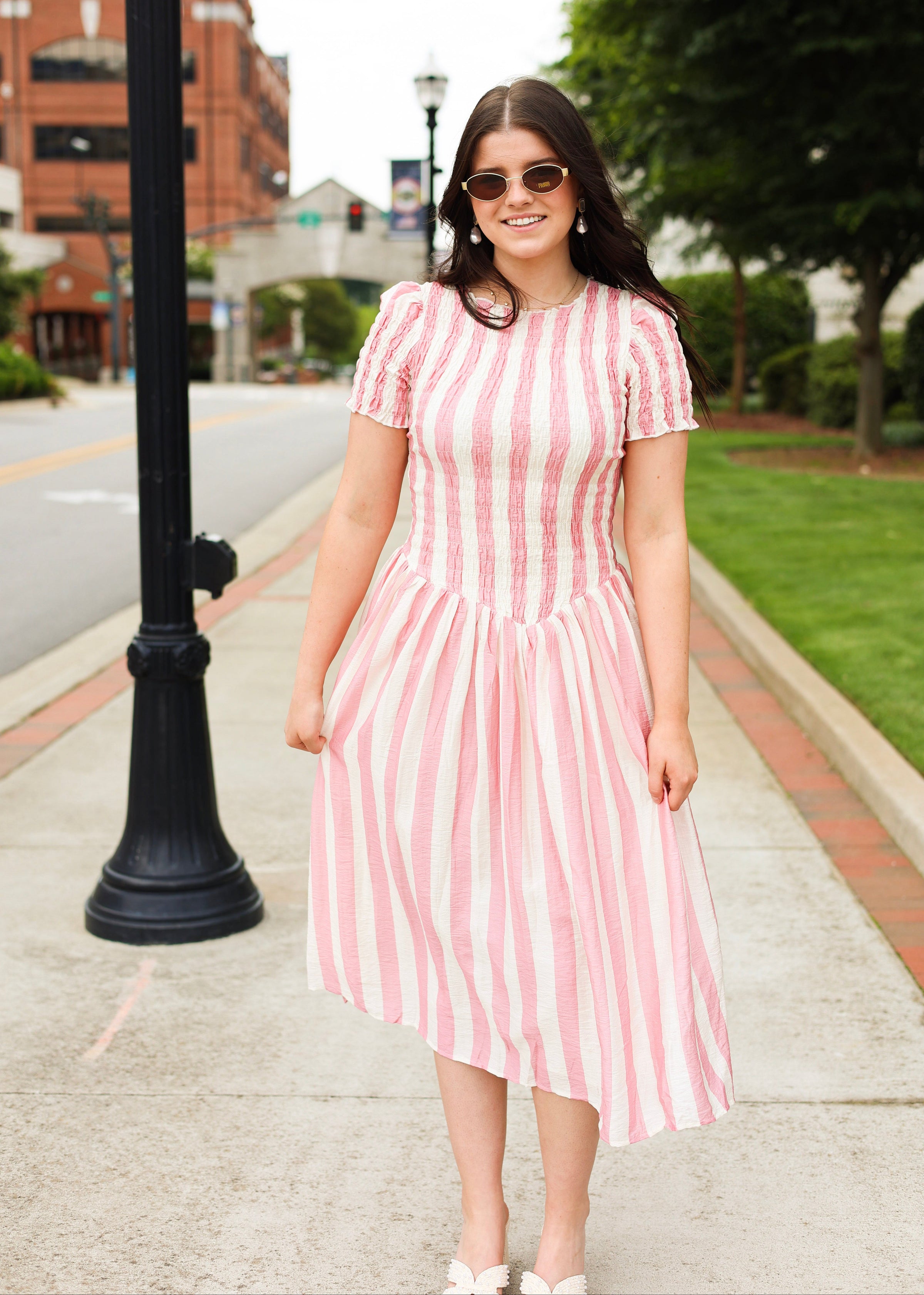 Woman in a pink and white striped dress standing on a sidewalk with buildings and trees in the background.